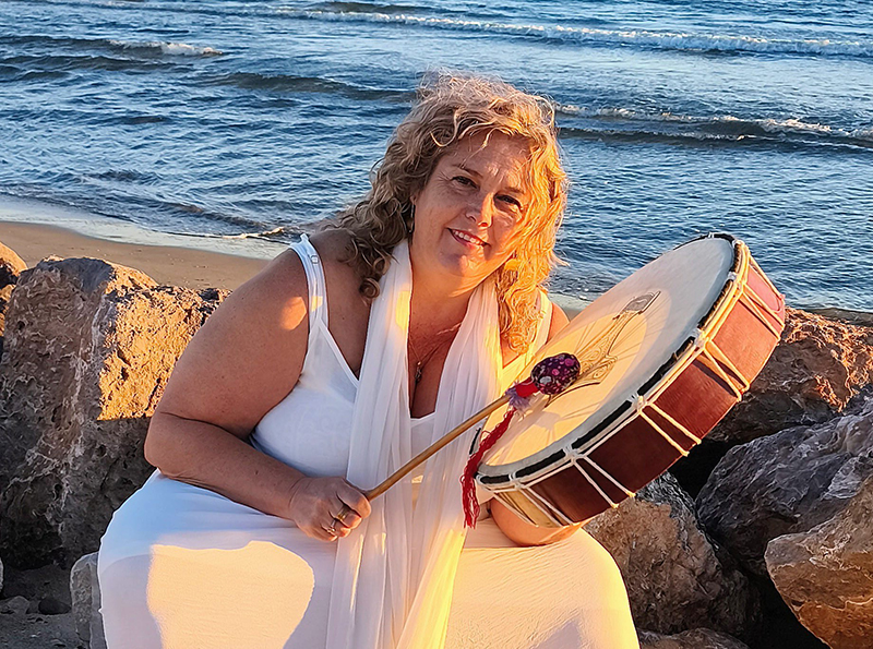 Retrato de Regina, sonriendo mientras sostiene y toca un tambor chamánico de piel en una playa al atardecer, sobre las rocas.