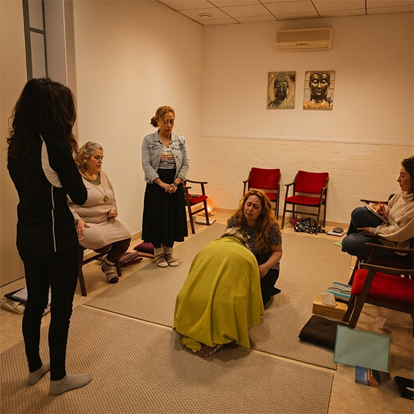 Sesión grupal de constelaciones familiares en un centro de terapia. Una mujer de pie guía a otra mujer arrodillada y cubierta con una manta amarilla en el centro, rodeadas por otros participantes observando.